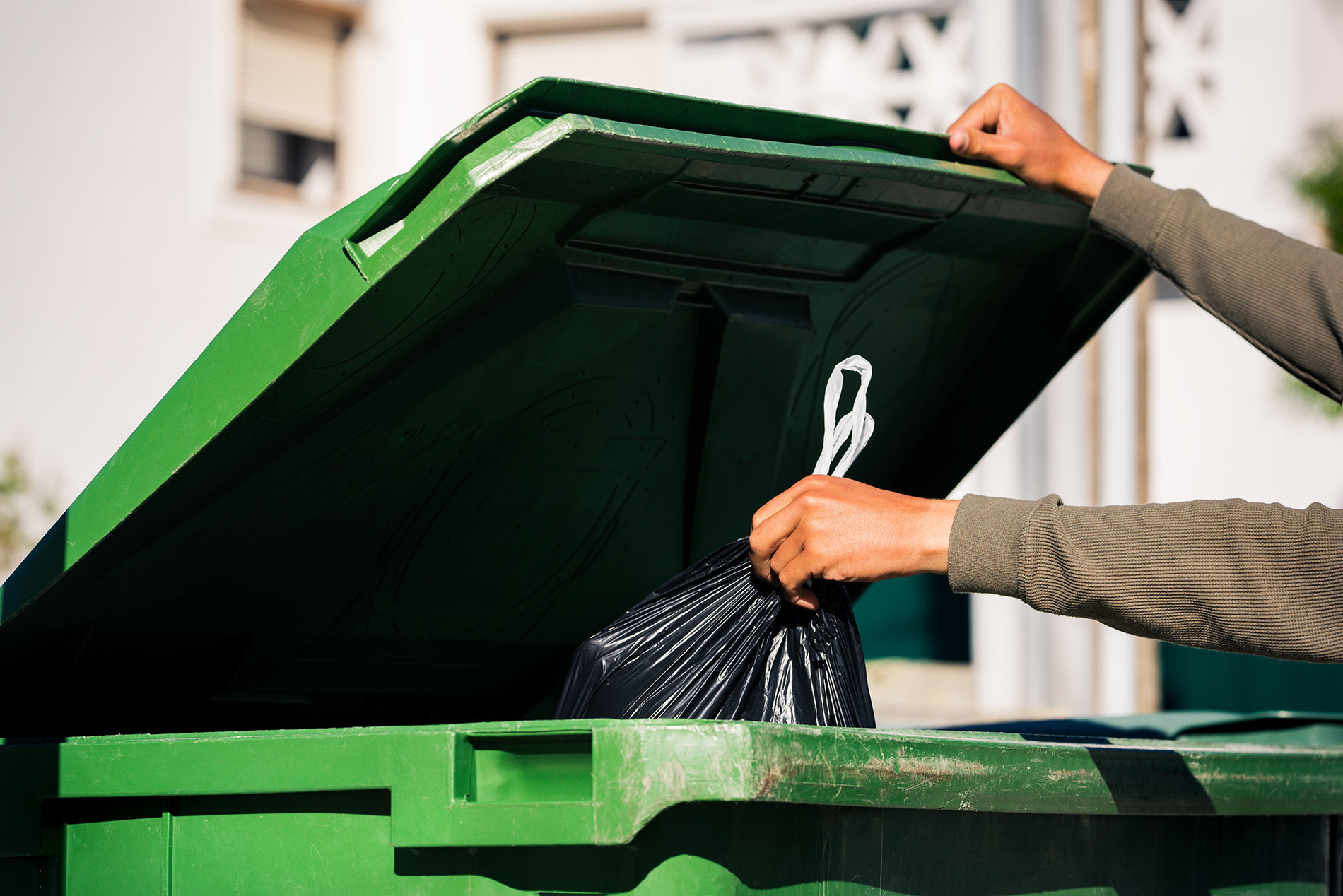 man throwing out black eco friendly recyclable trash bag in to big plastic green garbage container. take out the trash